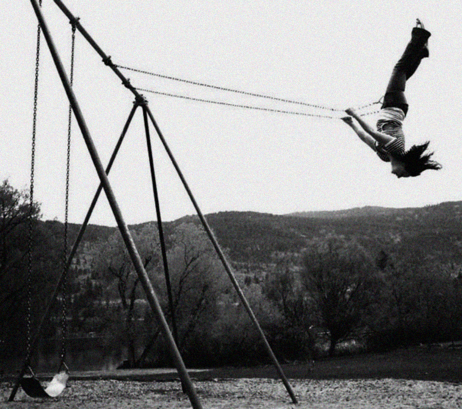 A person, pictured midair upside down, lets go of a swing set in a park. Trees and distant hills are visible in the background under a cloudy sky.