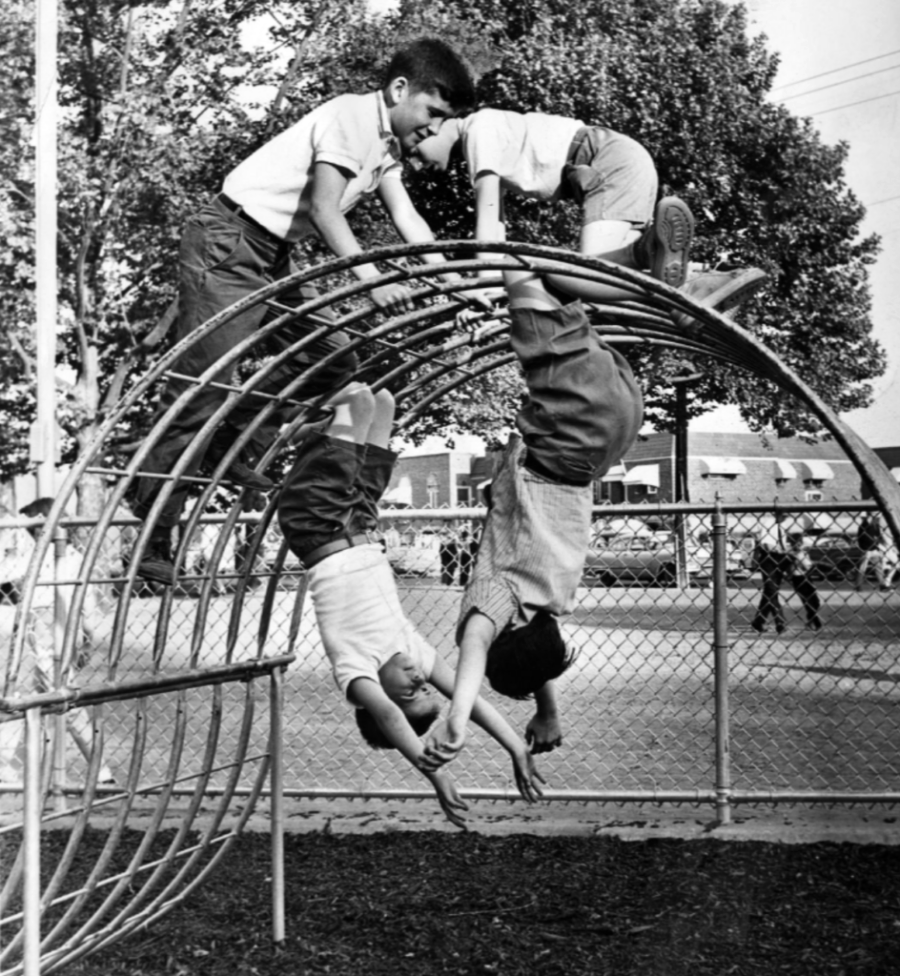 Four children play on a metal climbing dome at a playground, with two hanging upside down and two climbing on top. Trees, a chain-link fence, and houses are visible in the background.