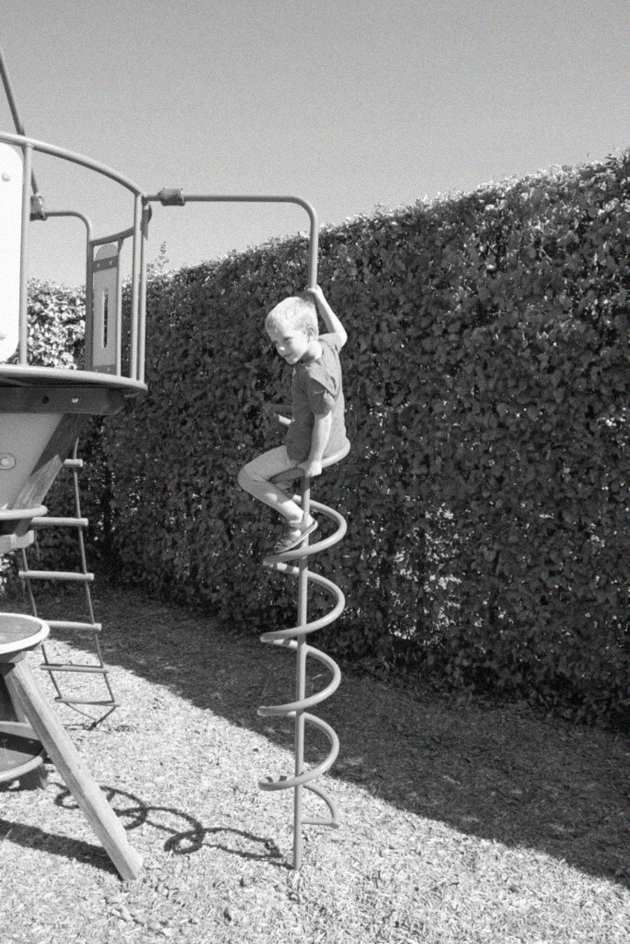 A child climbs a spiral ladder on a playground structure, holding onto the top bar. The scene is outdoors with a tall hedge in the background, and the ground is covered with wood chips. The photo is in black and white.