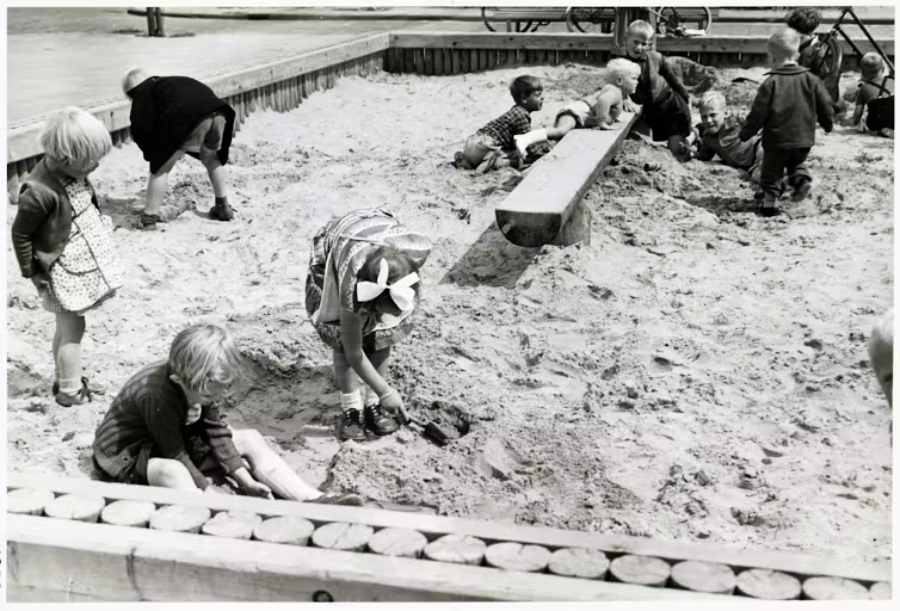 Several young children play in a large sandbox, digging and building with sand. Some sit or kneel, while others stand nearby. The scene is lively and outdoors on a sunny day.