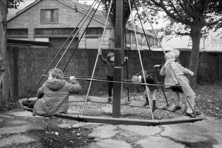 Four children play on a large, metal swing in an outdoor playground. Two hold onto the swing’s bars, while two others hang from the sides. The background shows trees, a fence, and an industrial-looking building.
