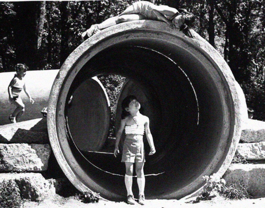 Three children play on large concrete pipes outdoors. One child lies on top of a pipe, another stands below inside the pipe looking up, and a third child climbs on a pipe in the background. Trees are visible behind them.
