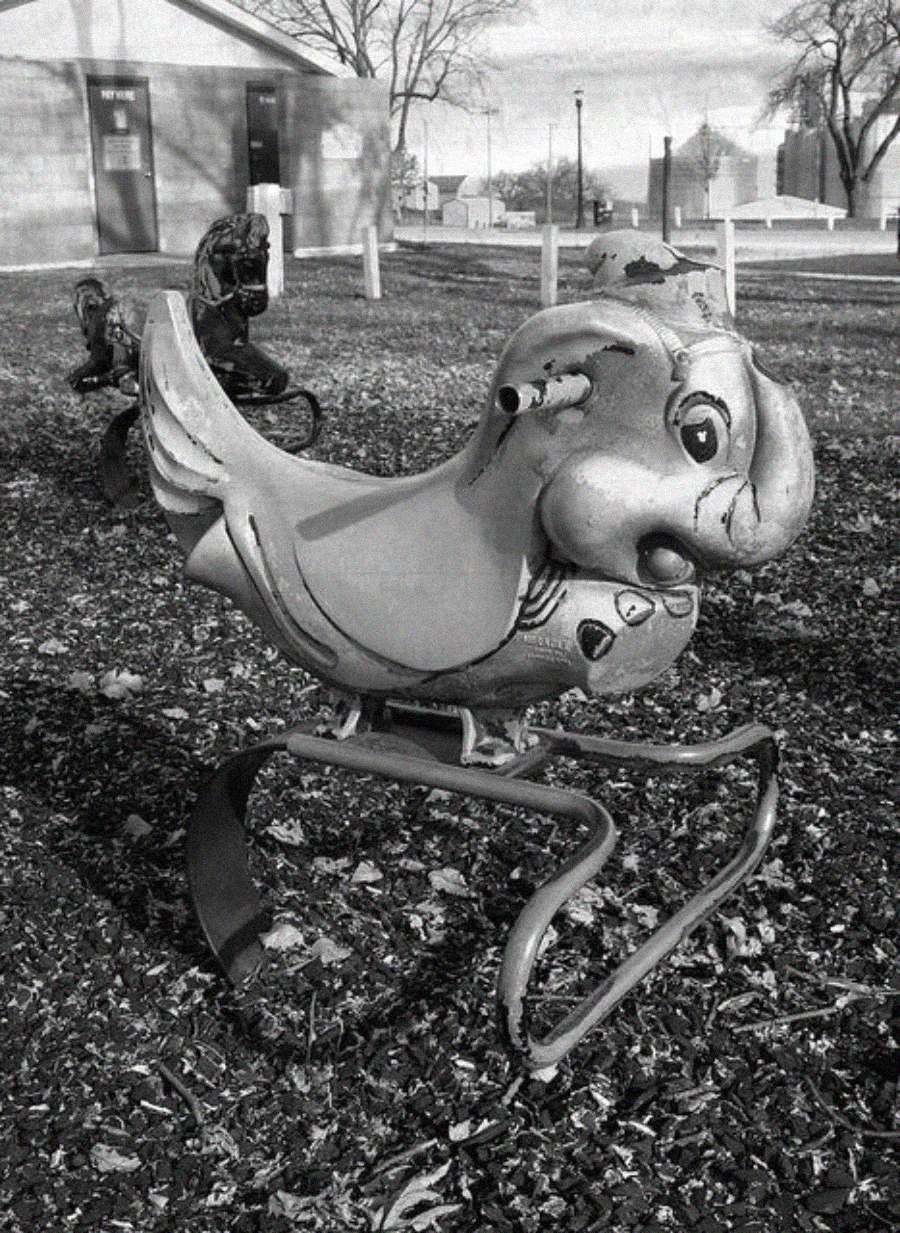 A playground spring rider shaped like a cheerful cartoon bird sits on mulch, with leafless trees and buildings in the background on a cloudy day.