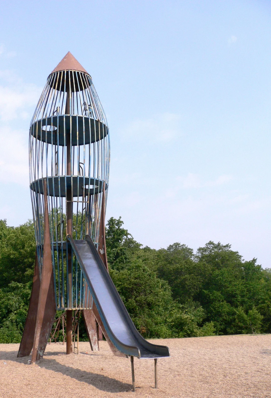 A tall, metal rocket-shaped playground structure with a slide stands on gravel in front of green trees and a clear blue sky.