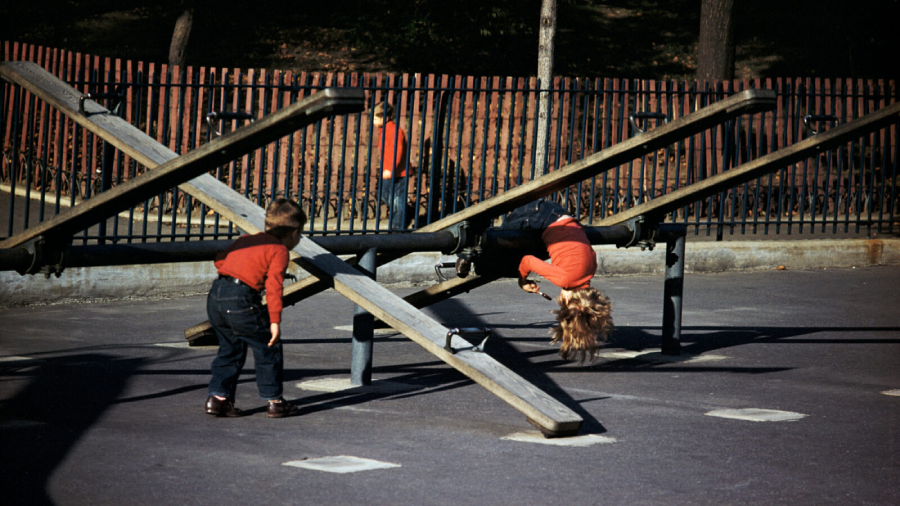 Two children in matching red shirts and dark pants play on wooden seesaws in a park. One child hangs upside down from a seesaw, while the other stands nearby. A fence and trees are in the background.