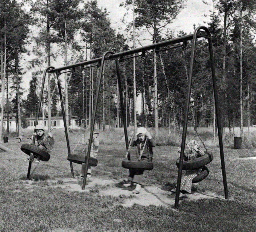 Three children sit on tire swings in a playground surrounded by tall trees. The children are wearing coats and hats, and the swings are made from repurposed tires hanging from a metal frame. The background shows grassy and wooded areas.