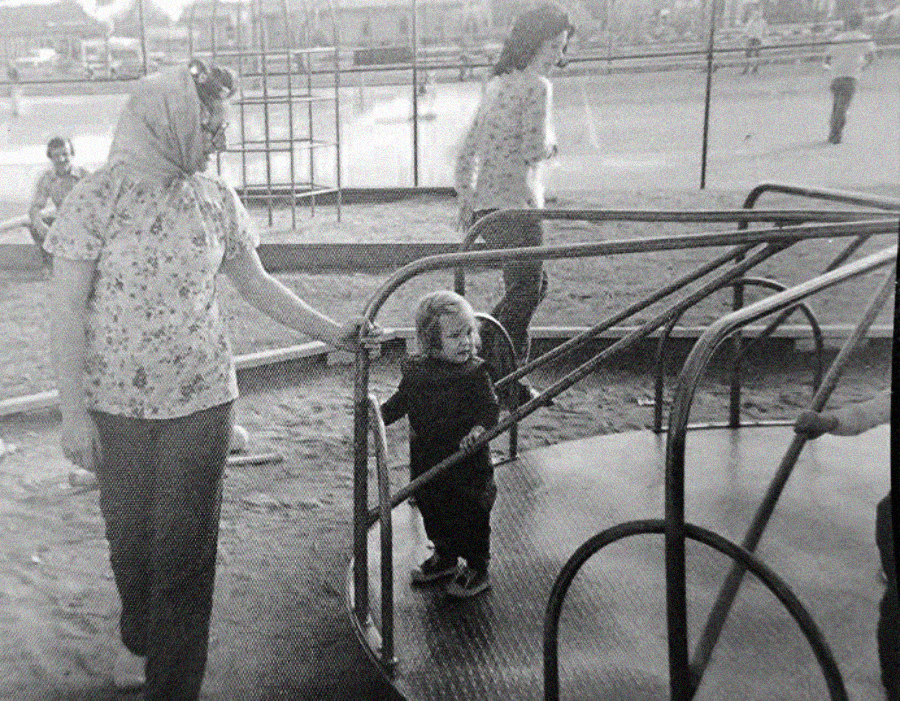 A black and white photo of a woman and a small child standing next to a playground merry-go-round, with other people in the background on a sandy playground.