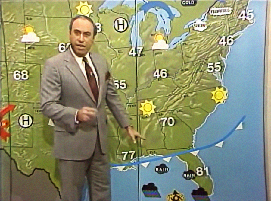 A weather forecaster in a suit stands in front of a U.S. weather map showing temperatures, weather icons, and a blue cold front line. The map displays mostly sun and varying temperatures across regions.