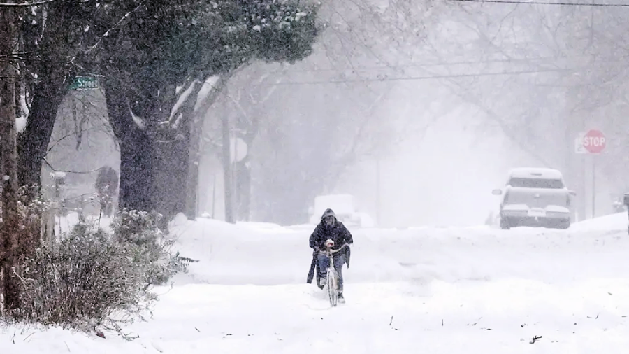 A person rides a bicycle through a snow-covered street during heavy snowfall; trees, parked cars, and street signs are visible under a blanket of snow.