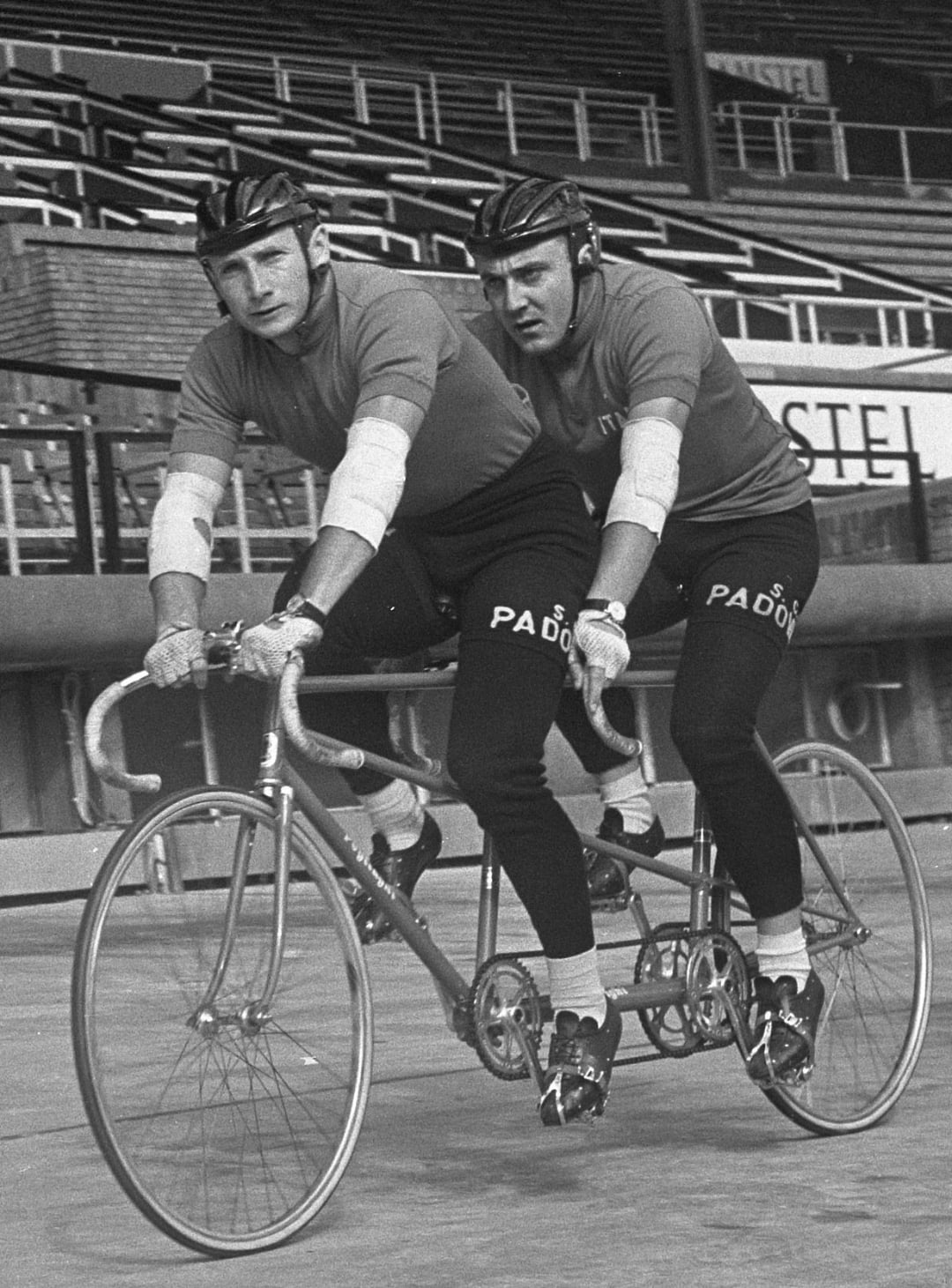Two male cyclists wearing matching outfits and helmets ride a tandem bicycle on an outdoor track. They both have bandaged elbows and are focused ahead, with empty stadium seating visible in the background.