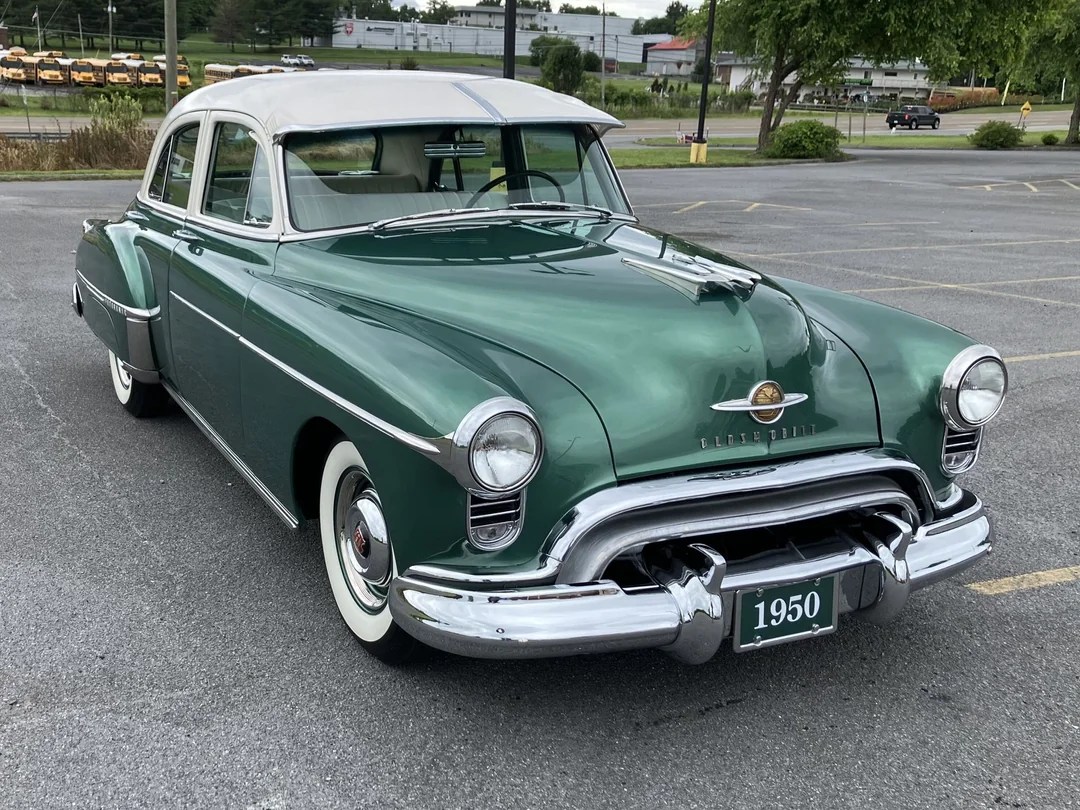 A vintage green 1950 Oldsmobile sedan with a white roof is parked in a nearly empty lot on an overcast day, showcasing its chrome trim and classic design.