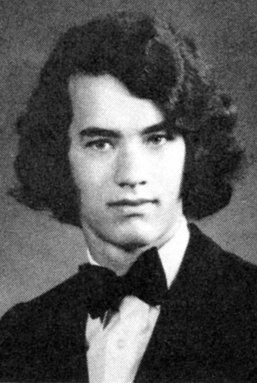 Black and white portrait of a young man with medium-length wavy hair, wearing a tuxedo jacket, white shirt, and bow tie, looking directly at the camera with a neutral expression.