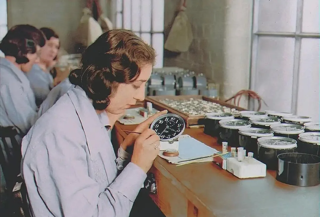 A woman in a light blue lab coat carefully paints numbers onto a clock face at a long workbench, surrounded by other women and numerous clock parts in a well-lit workshop.