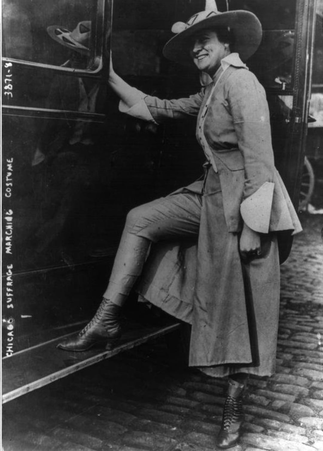A woman in early 20th-century clothing, including a long coat, wide-brimmed hat, and lace-up boots, stands with one foot on the step of a car. Handwritten text reads: "Chicago suffrage marching costume".