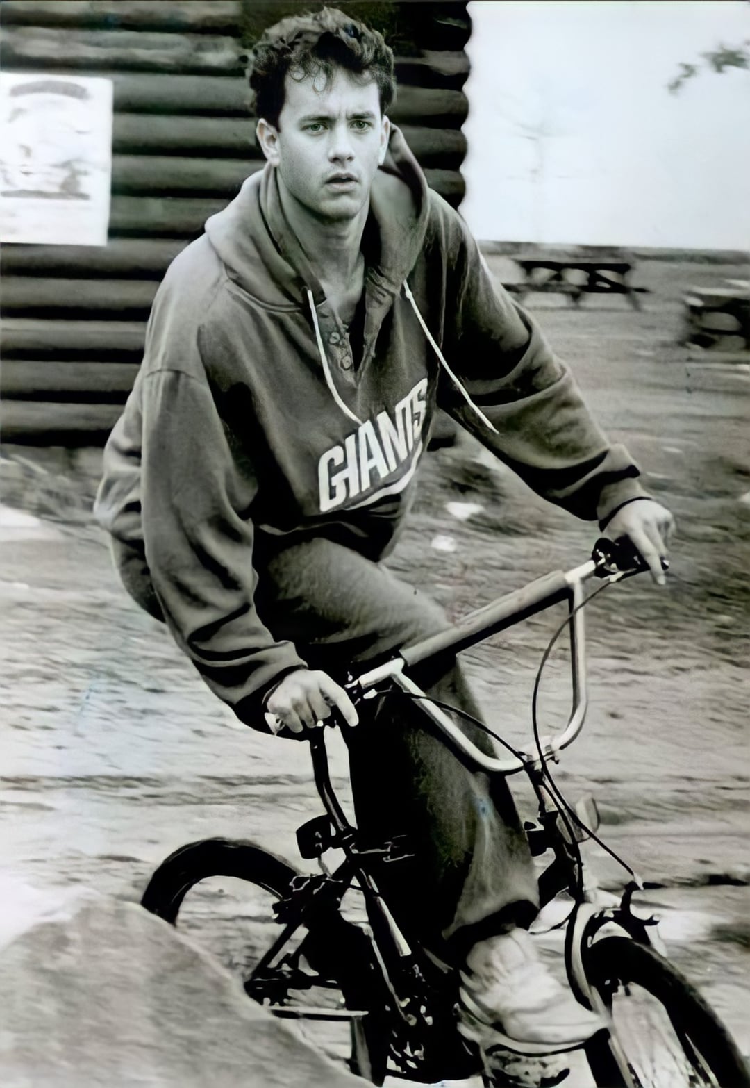 A young man wearing a "Giants" hoodie rides a BMX bicycle outdoors, looking intently ahead. The black-and-white photo shows a casual, active moment with blurred picnic tables and a log building in the background.