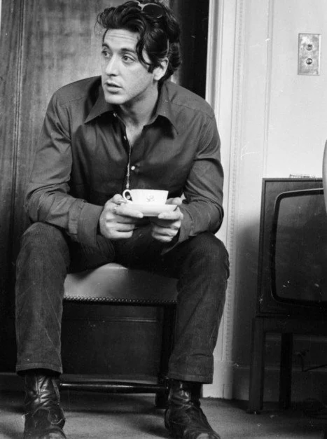 A man with dark, wavy hair sits on a chair holding a teacup and saucer, wearing a button-up shirt, dark pants, and boots. He looks to the side, with a vintage television and door in the background.