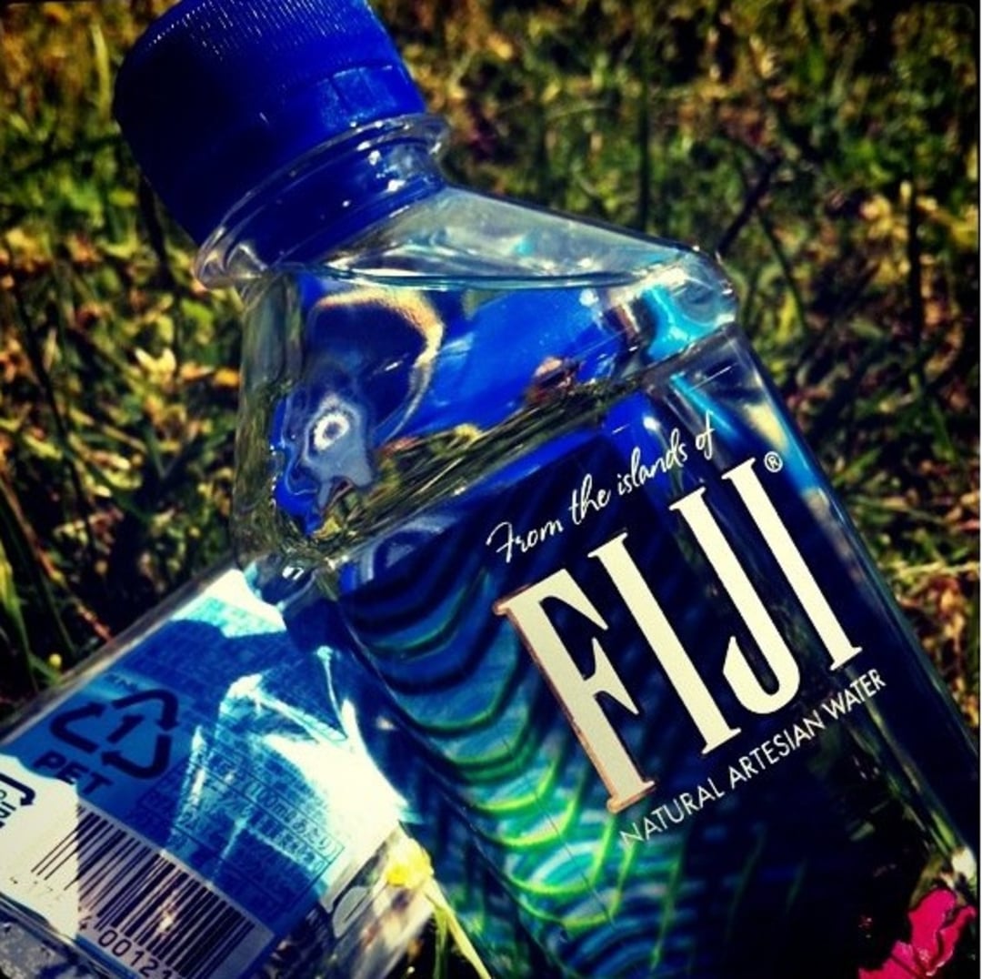 A close-up of the Fiji brands water bottle lying on grass in sunlight, with its blue cap on and a recycling symbol visible on a label in the background. The clear water and bottle reflect the blue sky.