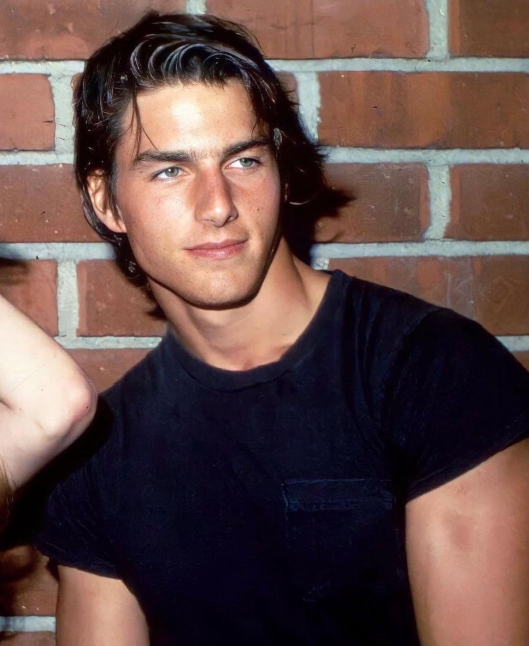 A young man with dark hair wearing a black t-shirt stands in front of a brick wall, looking slightly to the side with a calm expression.
