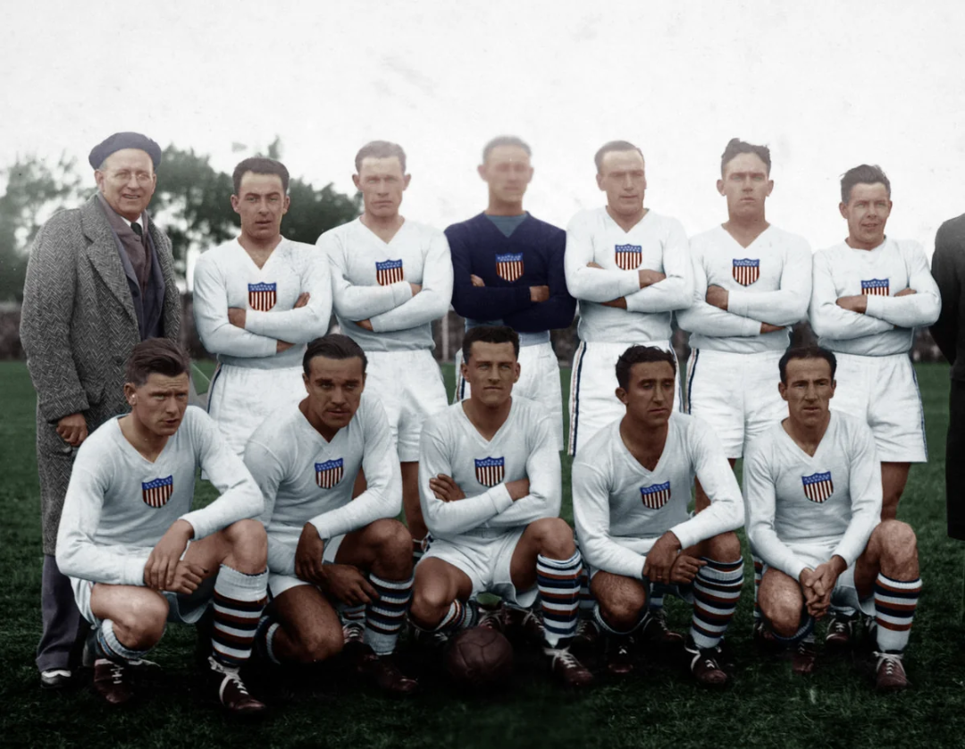 A vintage photo of a soccer team in white uniforms with USA crests, posing outdoors on grass; some players are standing with arms crossed, others are kneeling, with a coach in a beret on the left.