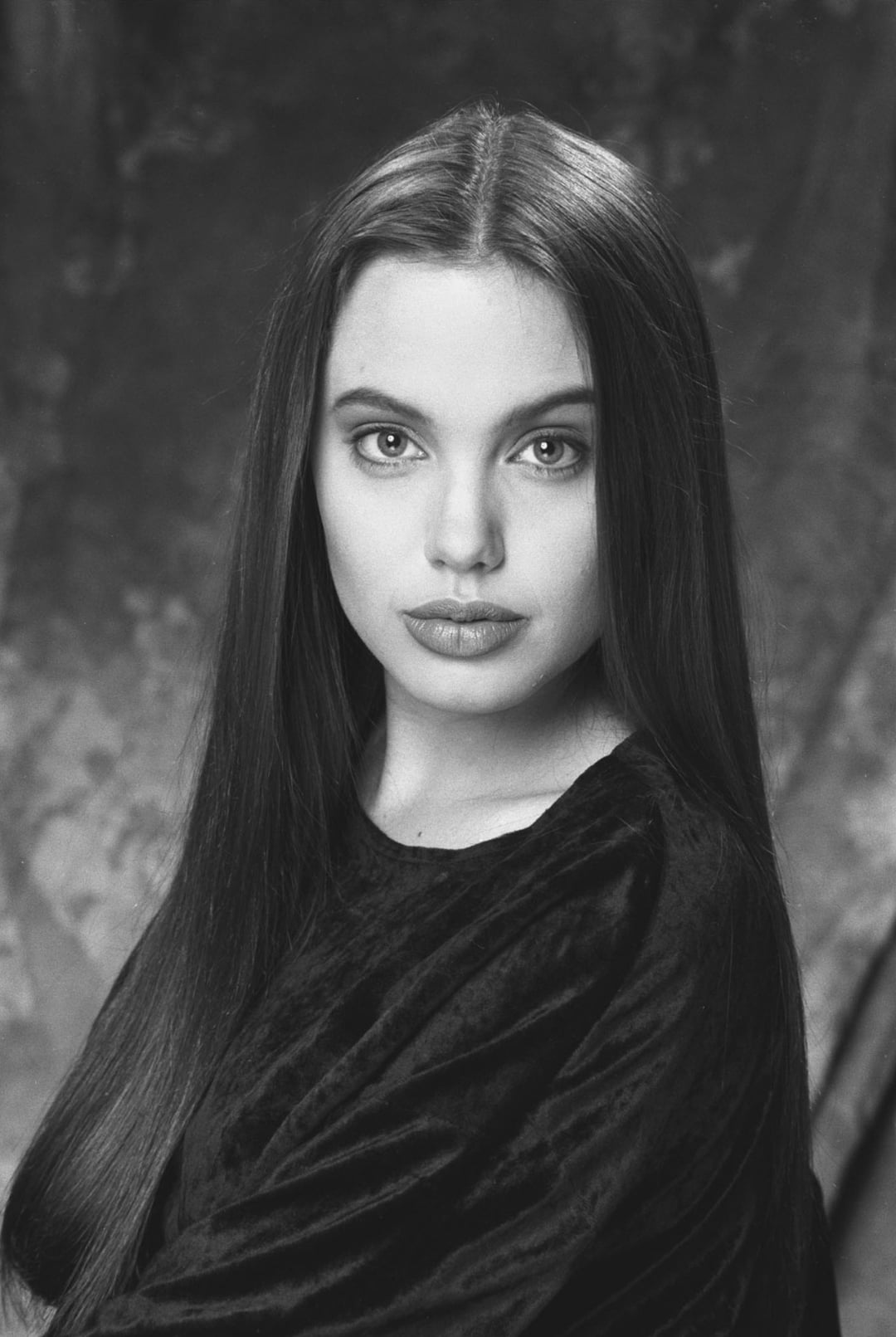 A young woman with long, straight dark hair and light eyes looks directly at the camera. She is wearing a dark velvet top and poses against a softly textured, dark background.