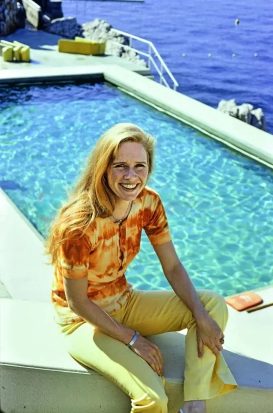 A woman with long blonde hair, wearing a tie-dye orange shirt and yellow pants, sits smiling on the edge of a pool with clear blue water. The sea and rocky coastline are visible in the background.