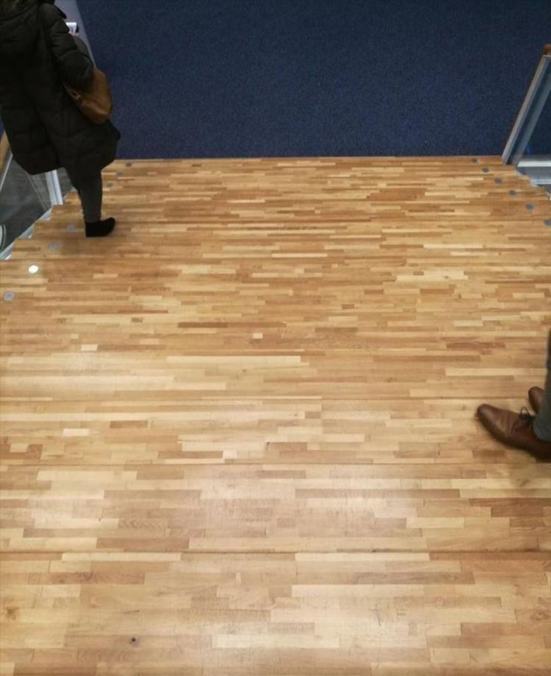 A pair of people are walking on what appears to be wooden flooring, but upon closer inspection, it’s actually stairs disguised by the uninterrupted wood pattern.