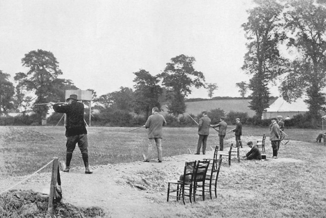 A black and white photo of several people standing in a row outdoors, shooting bows and arrows at targets. Empty chairs are lined up in the foreground; trees and grassy fields are visible in the background.