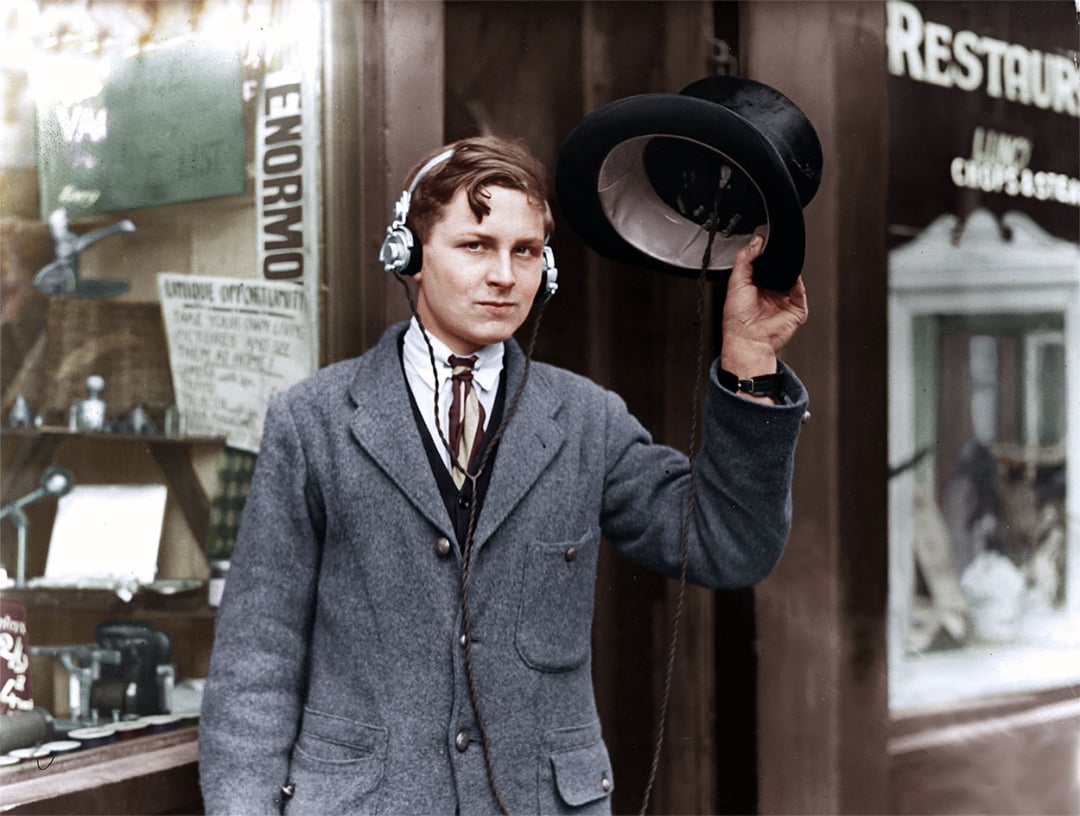 A young man stands outside a shop window, wearing early headphones and holding a hat with a radio receiver inside. He is dressed in a period suit and stands in front of a restaurant sign.