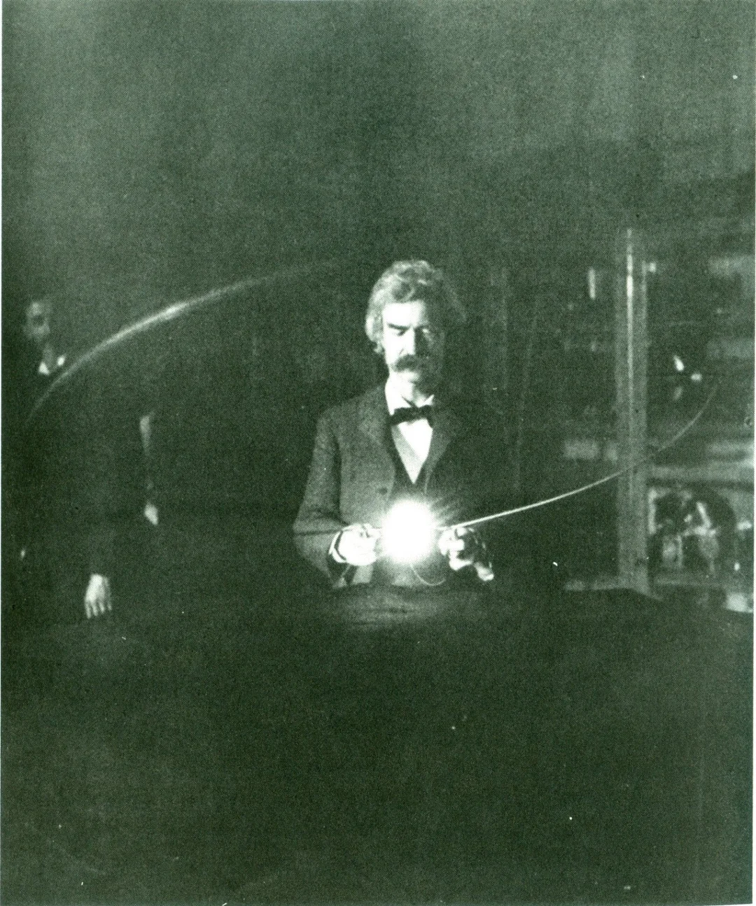 A man with a mustache in a suit stands in a dim room, holding a glowing object that emits bright light, with laboratory equipment and shadowy figures visible in the background.
