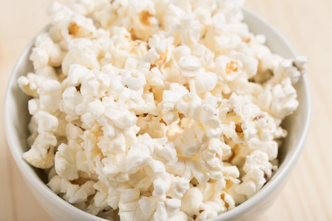 A white bowl filled with freshly popped popcorn sits on a light wooden surface. The popcorn appears fluffy and lightly salted, with some kernels slightly golden brown.