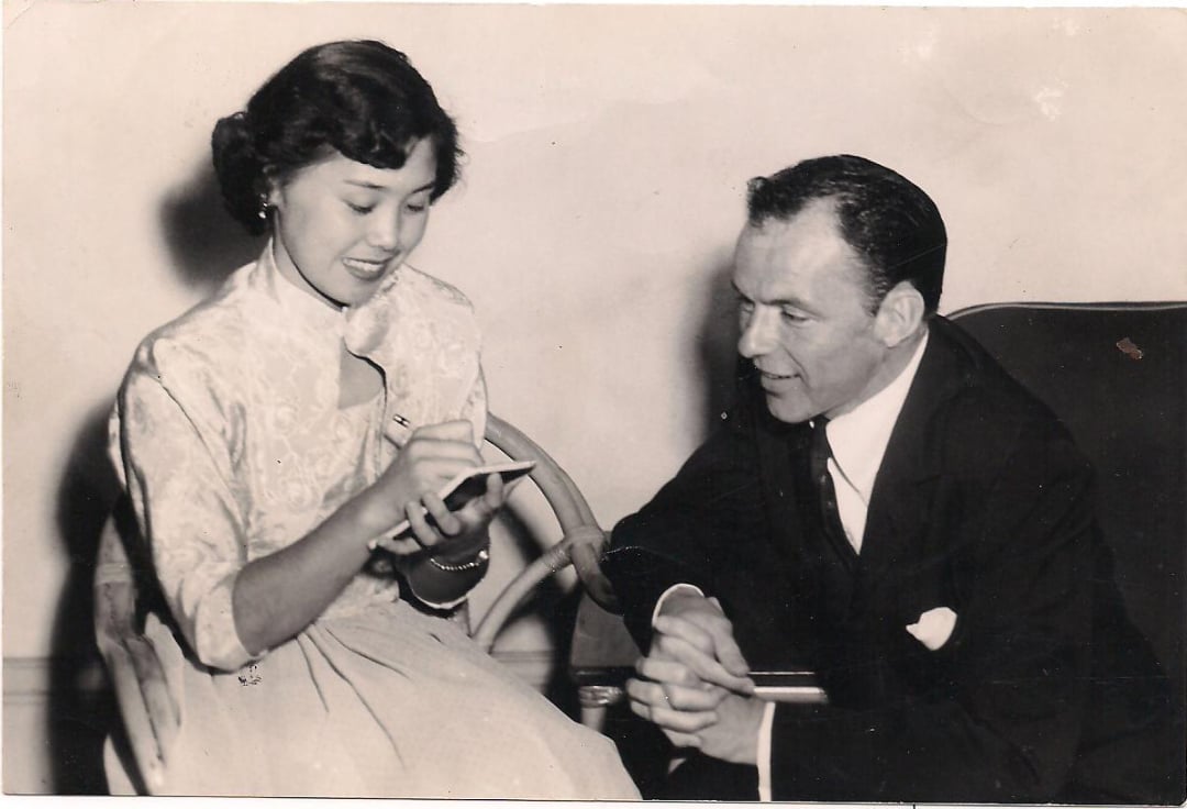A woman in a light-colored dress sits and writes on a notepad while smiling. A man in a dark suit sits beside her, leaning forward and looking at the notepad. Both appear engaged in conversation.