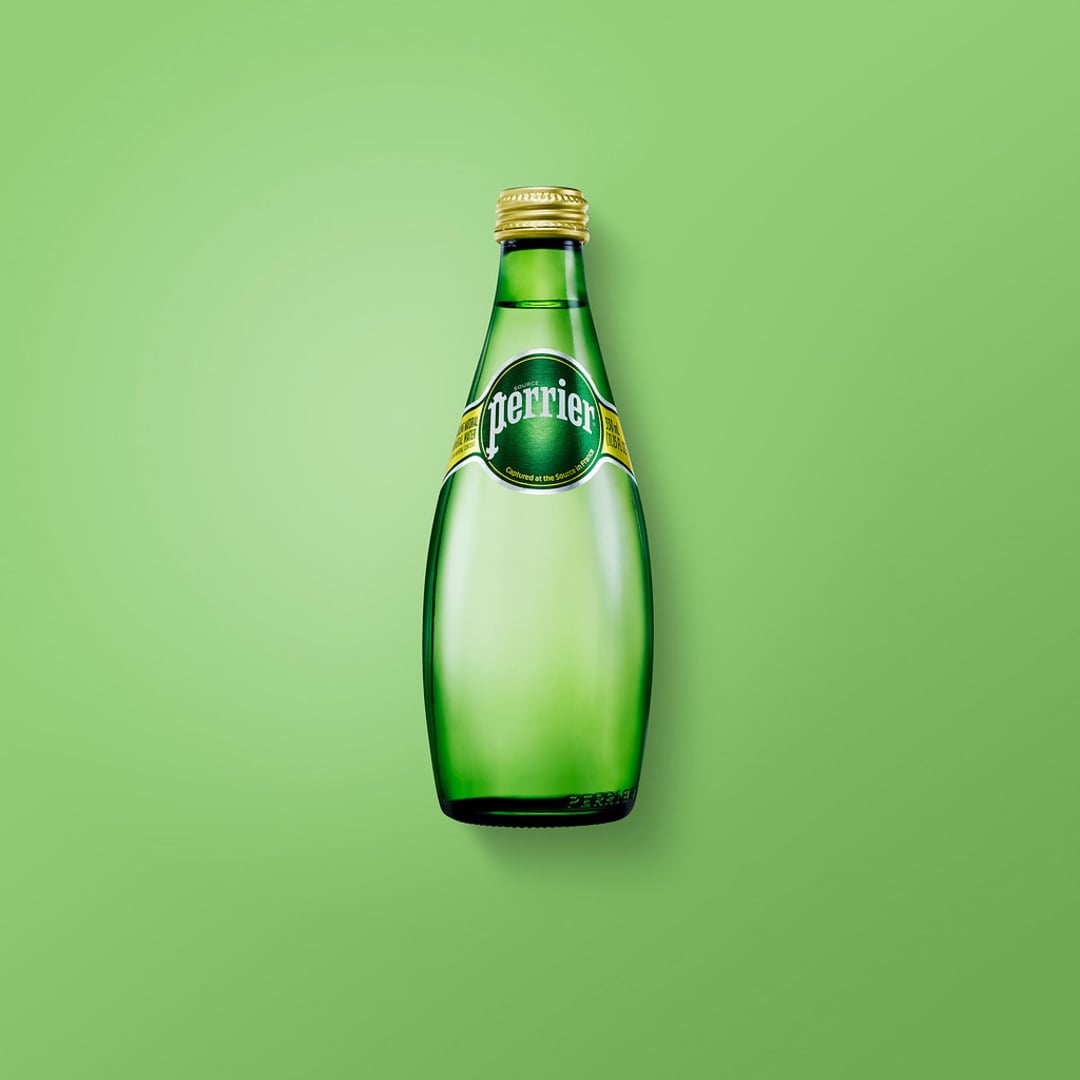 A green glass Perrier bottle with a gold cap is centered on a plain light green background. The bottle label is green with white and yellow accents.
