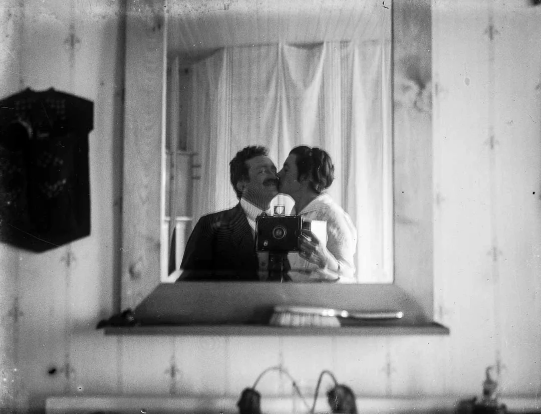 A black-and-white vintage photograph shows a man and woman taking a mirror selfie; the man holds an old-fashioned camera while the woman kisses him on the cheek. Their reflection appears above a mantle in a cozy room.