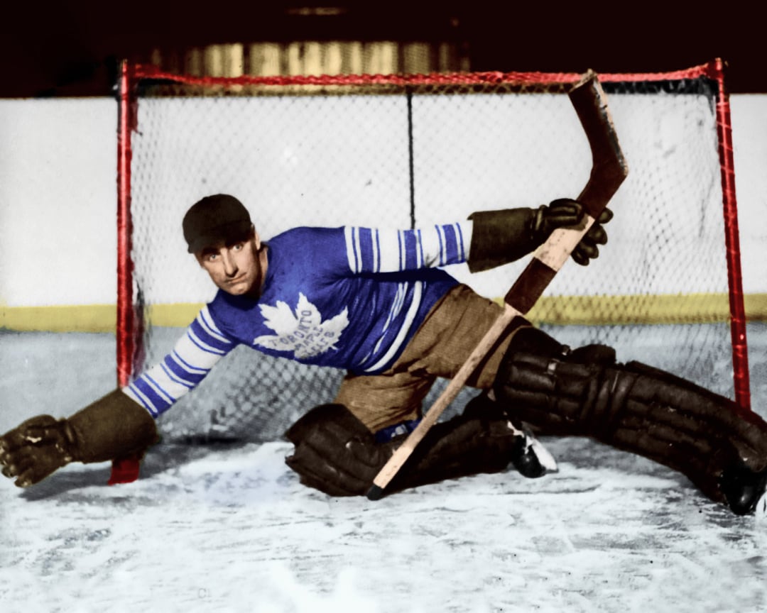 A vintage ice hockey goalie in old-fashioned gear and a blue Toronto Maple Leafs jersey makes a save in front of the net, stretching his leg and arms to block the puck.