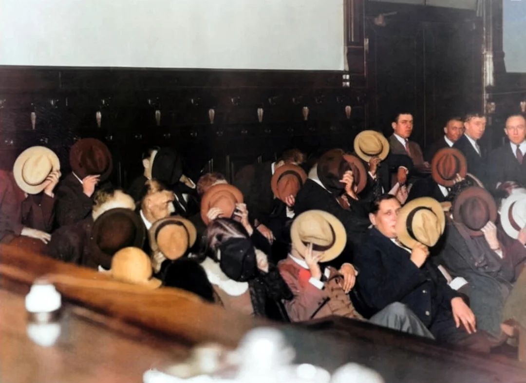 A group of people sitting in a courtroom cover their faces with hats, avoiding being identified; several men in suits stand in the background watching the scene.