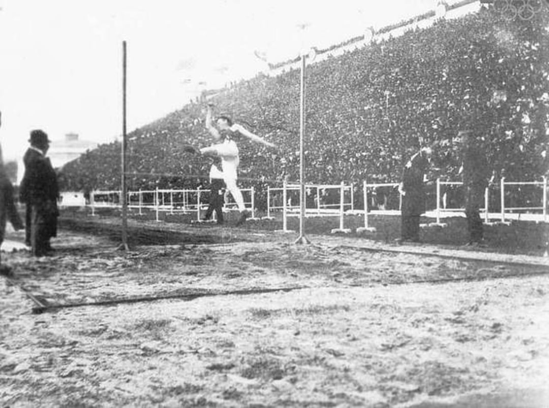 A black-and-white photo shows an athlete in mid-air attempting a high jump during a stadium event, with spectators filling the stands in the background and officials watching nearby.