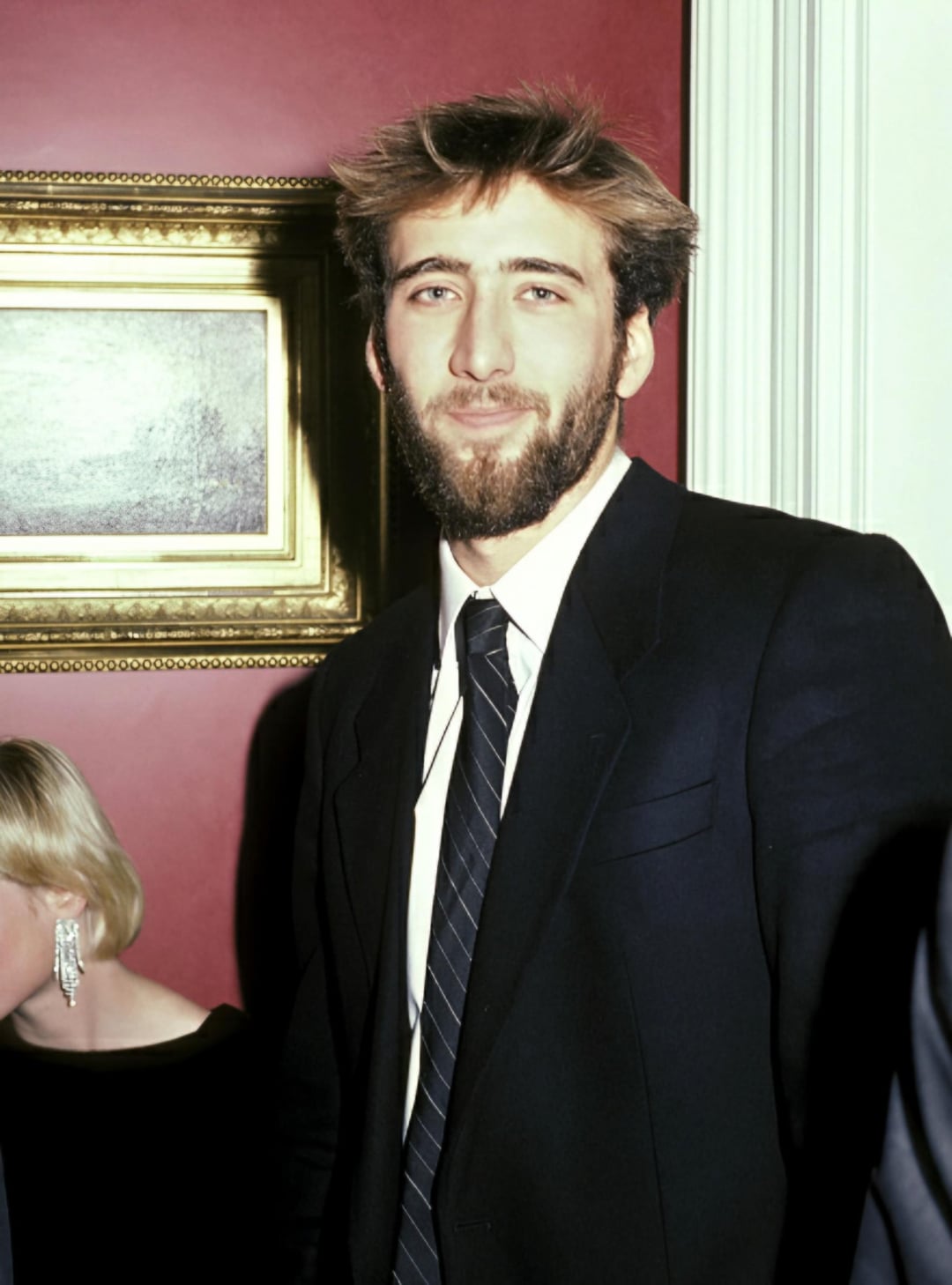 A man with tousled brown hair and a beard, wearing a dark suit, white shirt, and striped tie, stands in front of a red wall with a framed painting. A woman with short blonde hair and earrings is partially visible beside him.