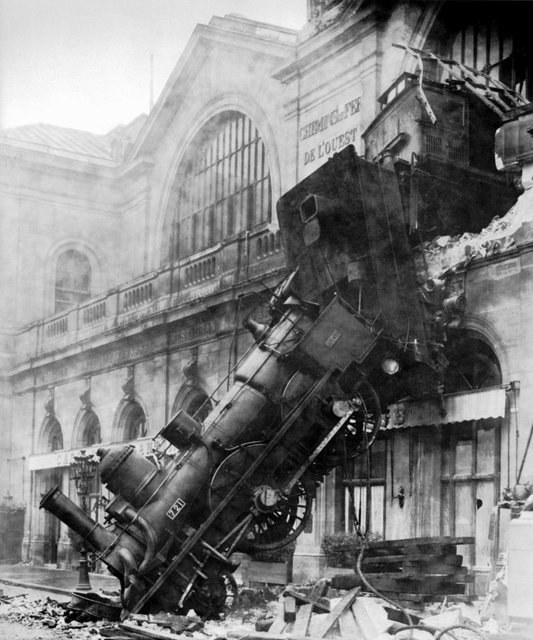 A black-and-white photo shows a steam locomotive that has crashed through the wall of a high train station, hanging vertically with its front end on the street below and debris scattered around.