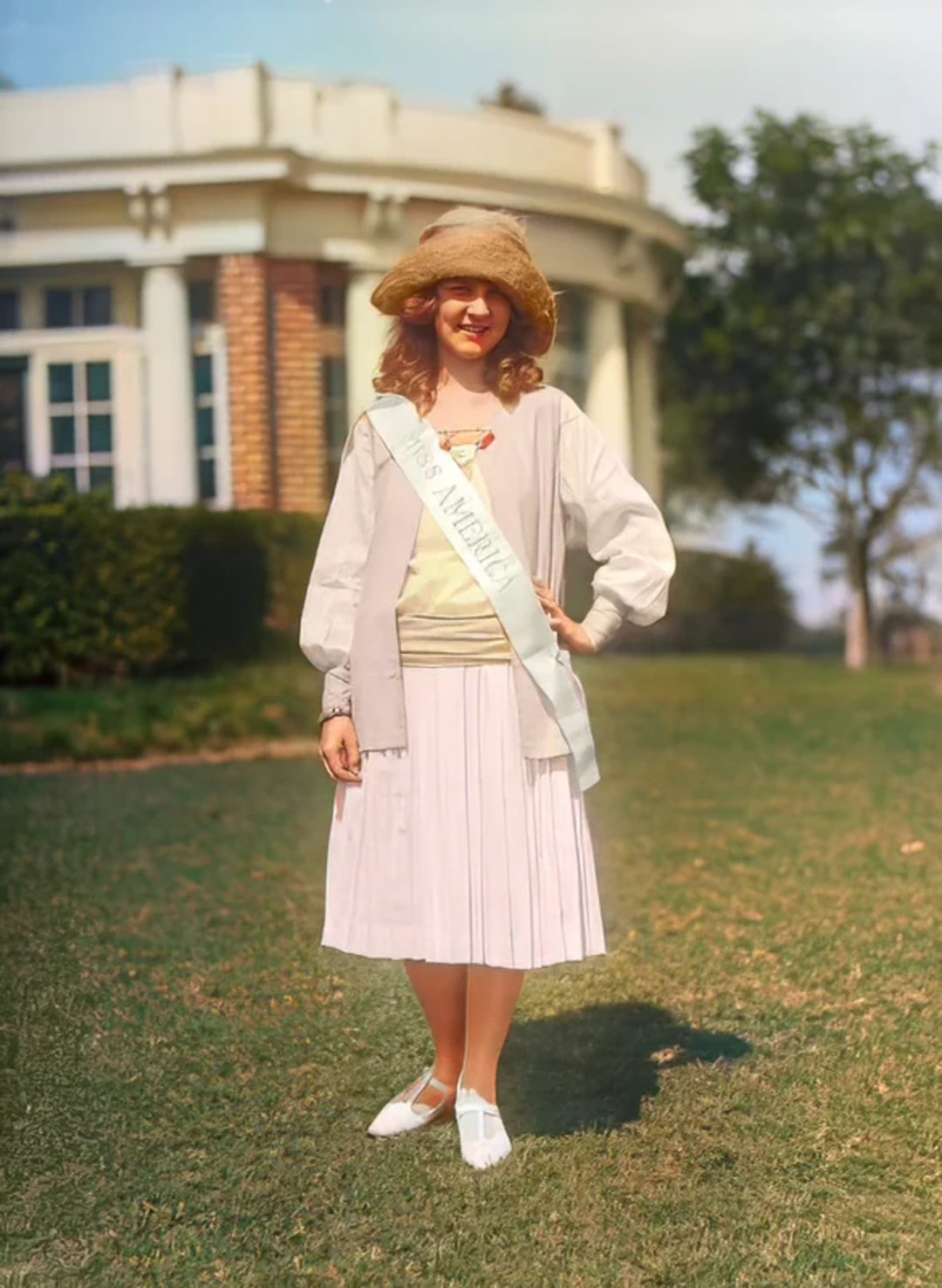 A woman stands on grass wearing a straw hat, light blouse, pleated skirt, and white shoes. She has a sash reading "Miss America." A large building and trees appear in the background under a clear sky.