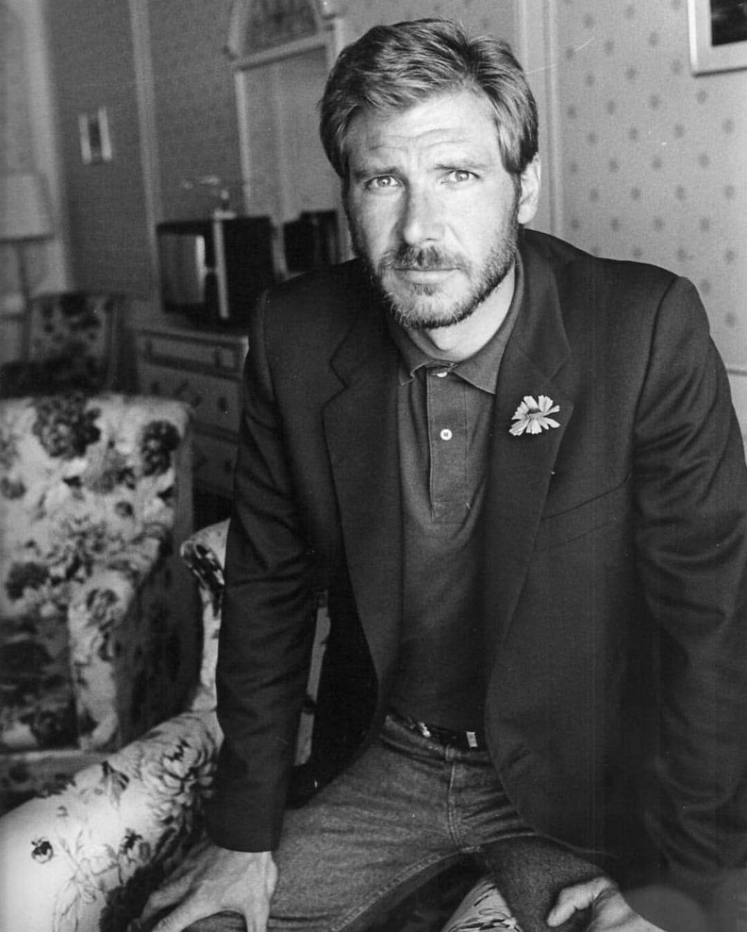 A man with light hair and a beard sits on the arm of a floral-patterned chair in a room with patterned wallpaper, wearing a blazer and jeans, looking into the camera with a calm expression.