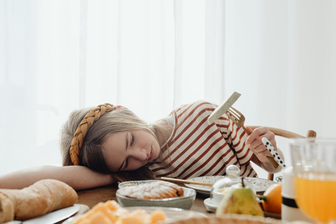 A young girl in a striped shirt rests her head on the table with her eyes closed, holding a fork and knife. Food, bread, and juice are on the table in front of her. She appears tired or sleepy.