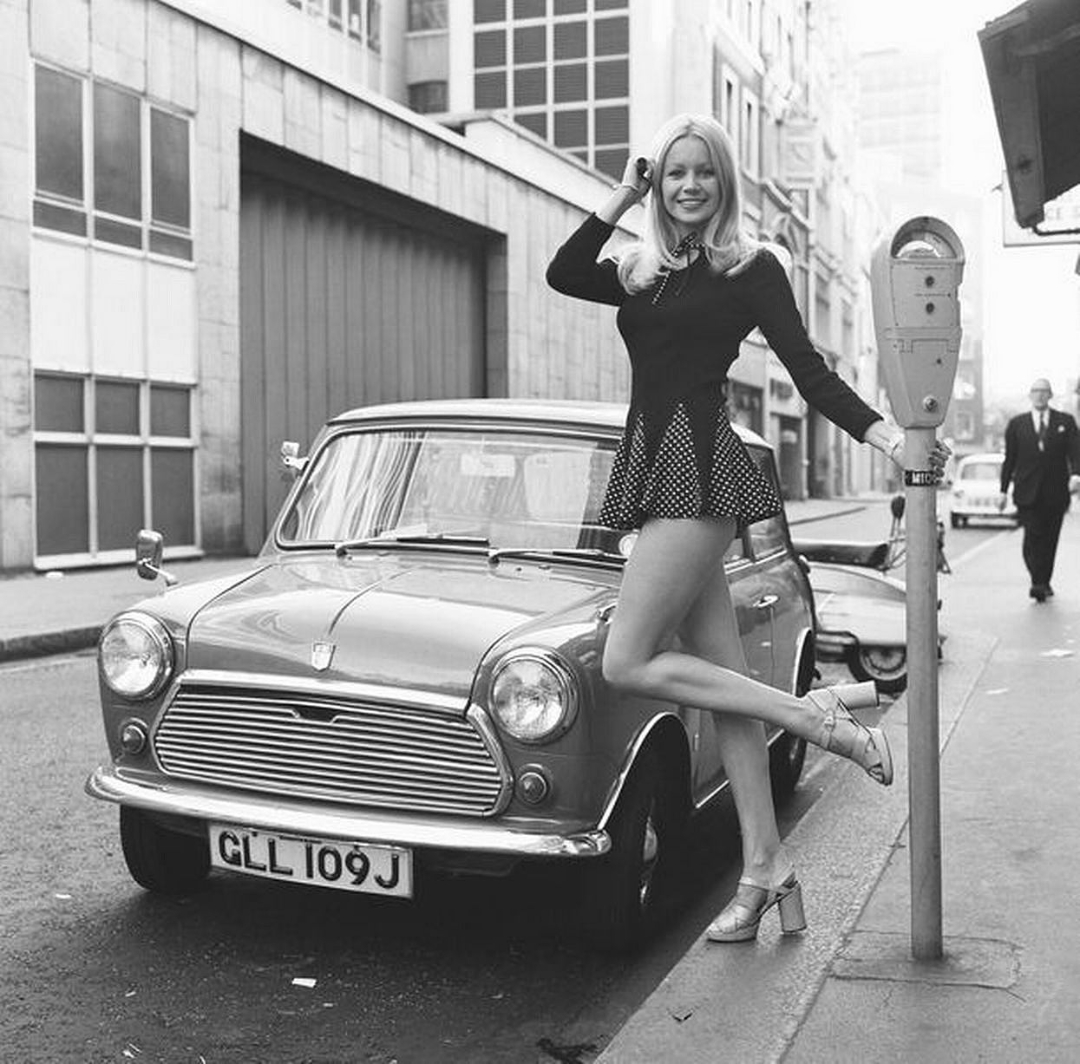 A woman in a short skirt and heeled sandals stands by a parking meter next to a classic Mini car on a city street, smiling and posing with one hand on her hip, in a vintage black-and-white photo.