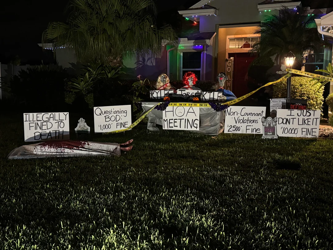 A yard at night is decorated with Halloween props, fake bodies, and tombstones. Signs read messages like “Illegally fined to death,” “Questioning BOD: $1,000 fine,” and “HOA meeting.” Yellow caution tape surrounds the area.