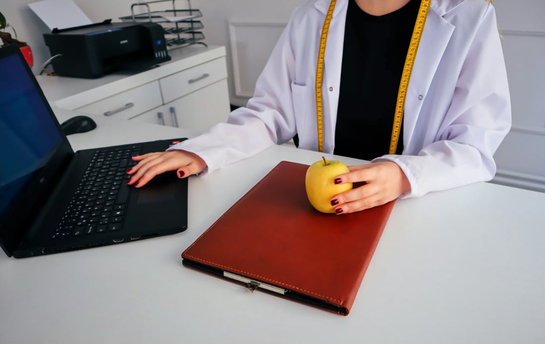 A person in a white lab coat sits at a desk using a laptop, with a yellow apple and a closed brown folder in front of them. A yellow measuring tape hangs around their neck.