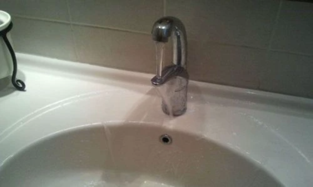 A bathroom sink with a faucet positioned too far back, causing the water stream to barely reach the edge of the sink instead of flowing directly into the basin.