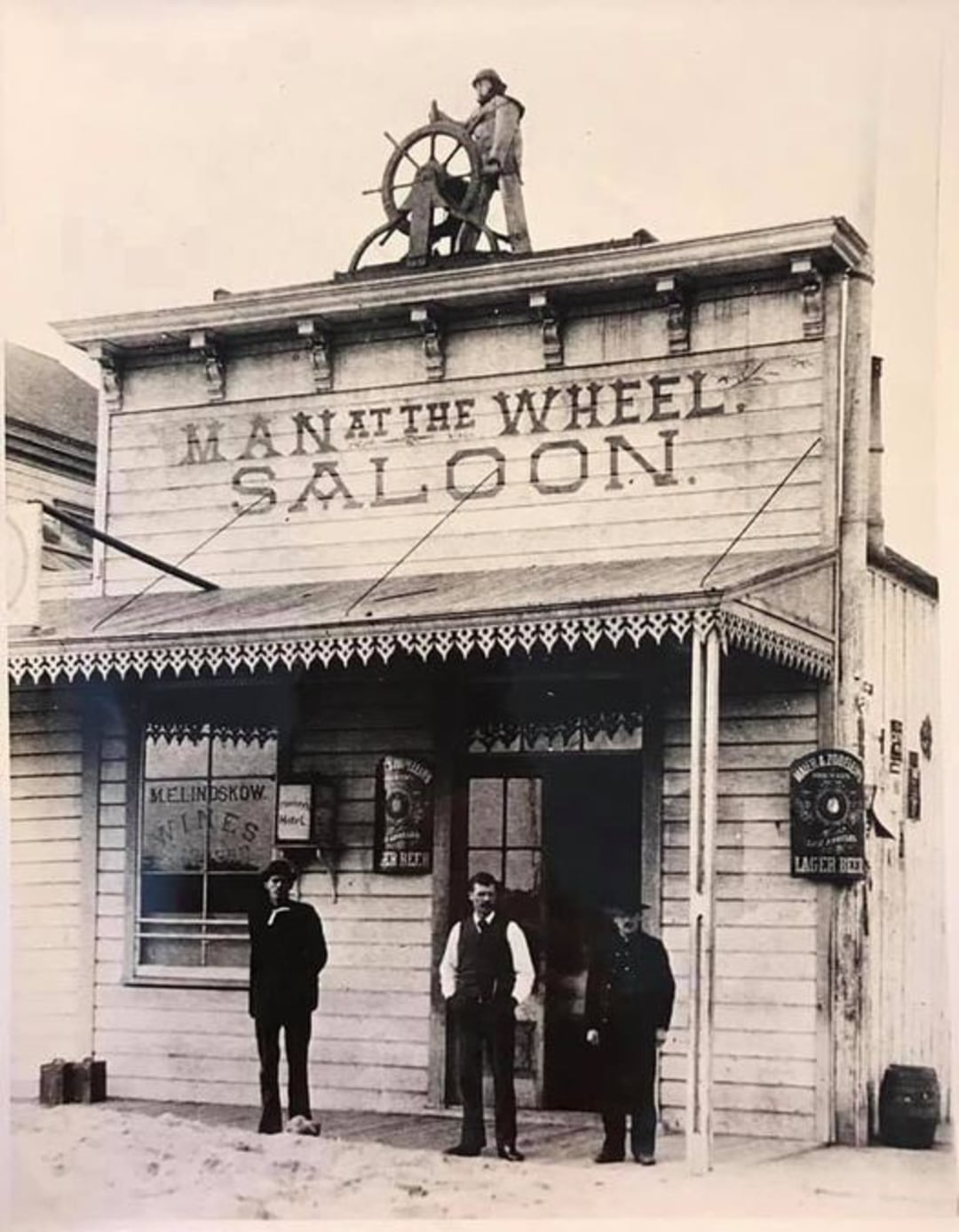 Black-and-white photo of four men in front of a wooden building labeled "Man at the Wheel Saloon." One man stands on the roof behind a ship’s wheel; three others stand on the ground in front of the entrance.