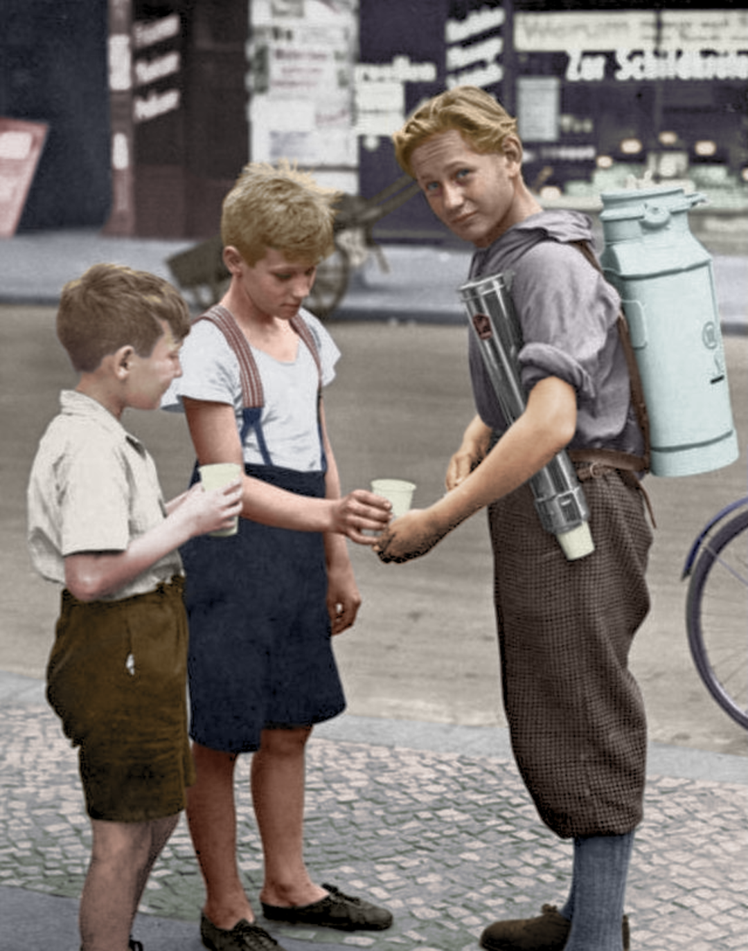 Three boys stand on a city sidewalk; one boy, carrying a large milk canister on his back, pours milk into cups held by the other two boys. The street and shop signs are visible in the background.