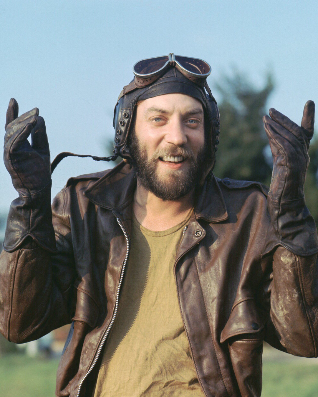 A man with a beard wears a leather aviator jacket, gloves, and vintage pilot goggles on his head, smiling outdoors with his hands raised and trees blurred in the background.