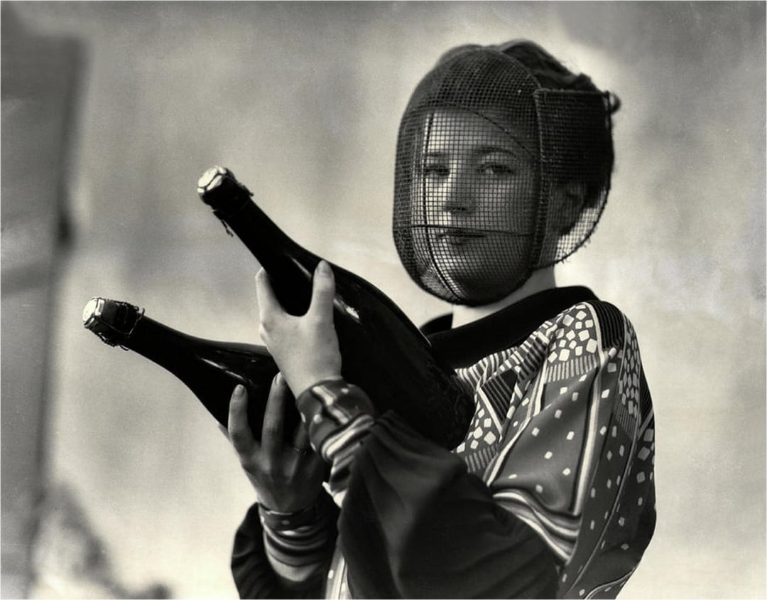 A woman in patterned clothing and a mesh face veil holds two large champagne bottles, looking at the camera with a slight smile. The photo is black and white, with a soft-focus background.