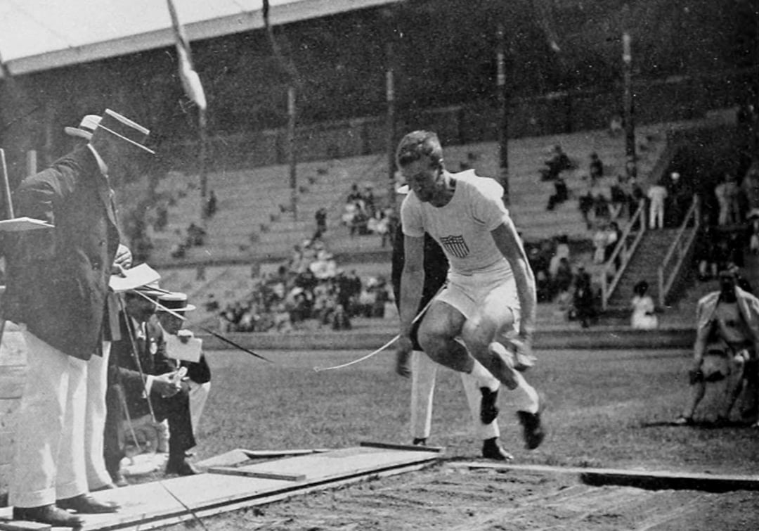 Black and white photo of a long jumper in mid-air at a stadium, wearing a USA shirt. An official records the jump while spectators watch from the stands in the background.
